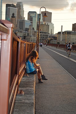a girl sits on a bridge with a city in the background.