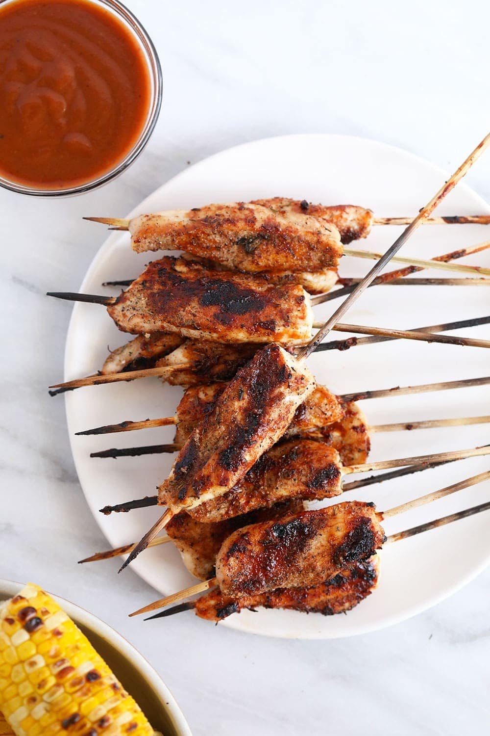 These BBQ Chicken Skewers are the perfect summer meal! Grab some skewers, BBQ sauce, and your favorite BBQ side dish and let's grill! Grilled skewers on plate next to bbq sauce and grilled corn.