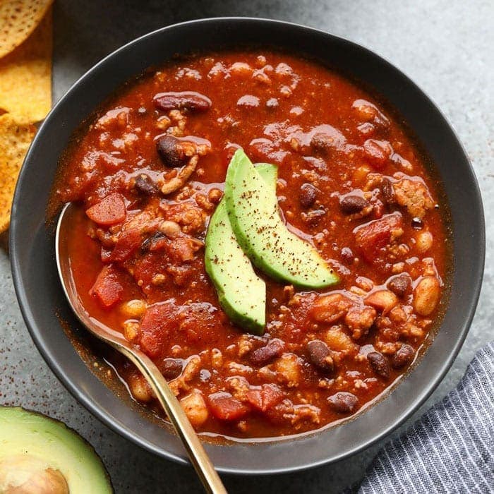 a bowl of turkey chili with avocado and tortilla chips.