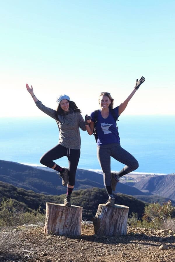 Two women from Wellfit Malibu standing triumphantly on top of a tree stump, overlooking the ocean.