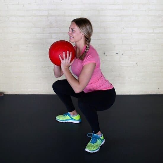 A woman performs a full-body AMRAP exercise while squatting down with a red ball.