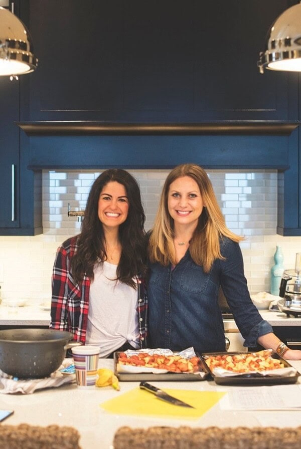 two women standing in front of a kitchen with pizzas on the counter.