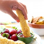 A person is dipping coconut-crusted chicken fingers into a bowl of strawberries.