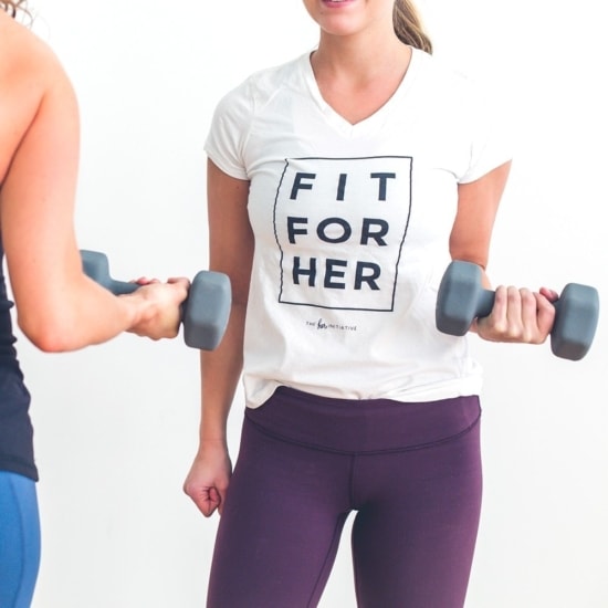 Two women from Fit For Her Minneapolis holding dumbbells wearing a white t-shirt.