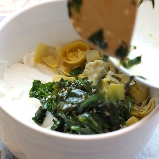 a bowl full of greens and cheese in a white bowl, Lightened Up Spinach and Artichoke Dip.