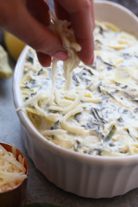 A person artfully drizzling cheese on a dish of Lightened Up Spinach and Artichoke Dip.