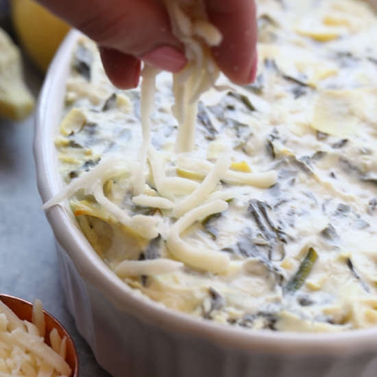A person artfully drizzling cheese on a dish of Lightened Up Spinach and Artichoke Dip.