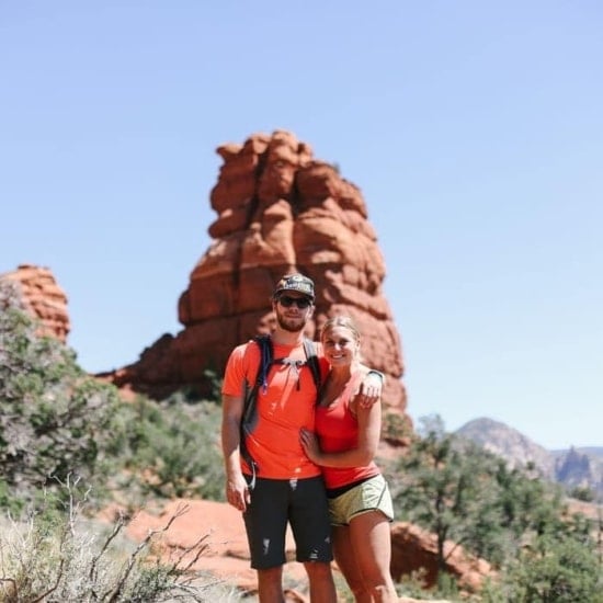 A couple on a surprise trip posing in front of a red rock formation.