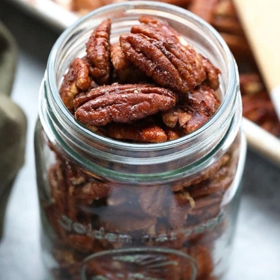 Roasted pecans displayed in a glass jar with a wooden spoon.