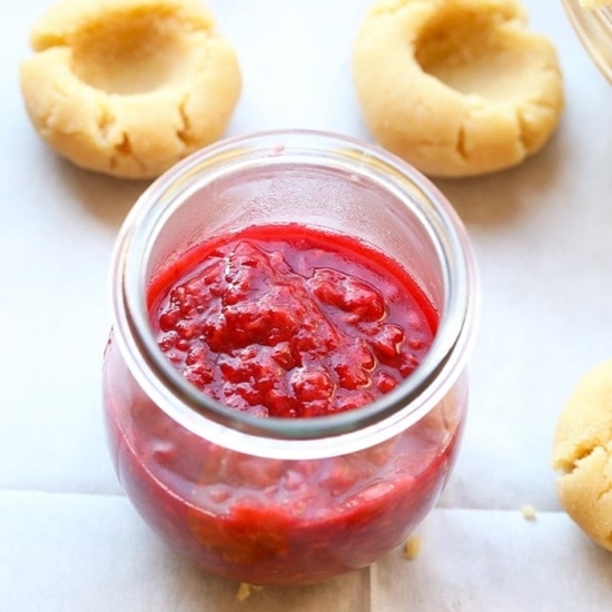 A jar of raspberry jam beside Raspberry Thumbprint Cookies on a baking sheet.