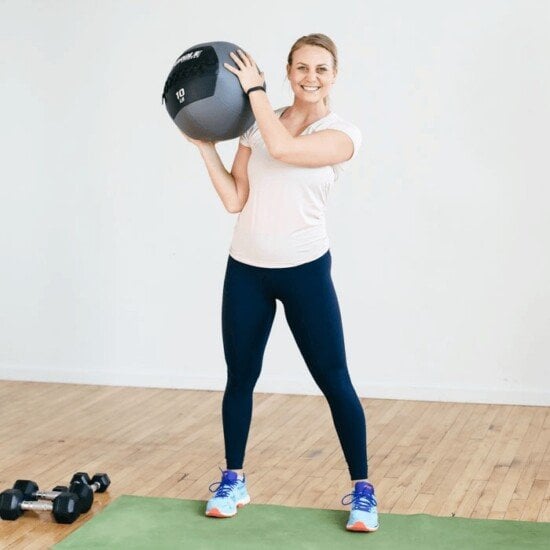 A woman demonstrating a plyo workout with an exercise ball against a white wall.