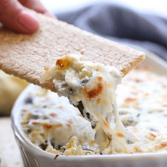 A person is dipping a cracker into a bowl of Lightened Up Spinach and Artichoke Dip.