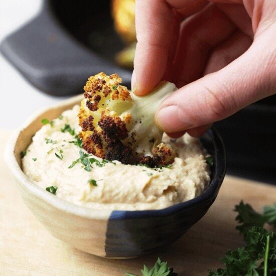 A person dunking a piece of roasted cauliflower into a bowl of hummus.