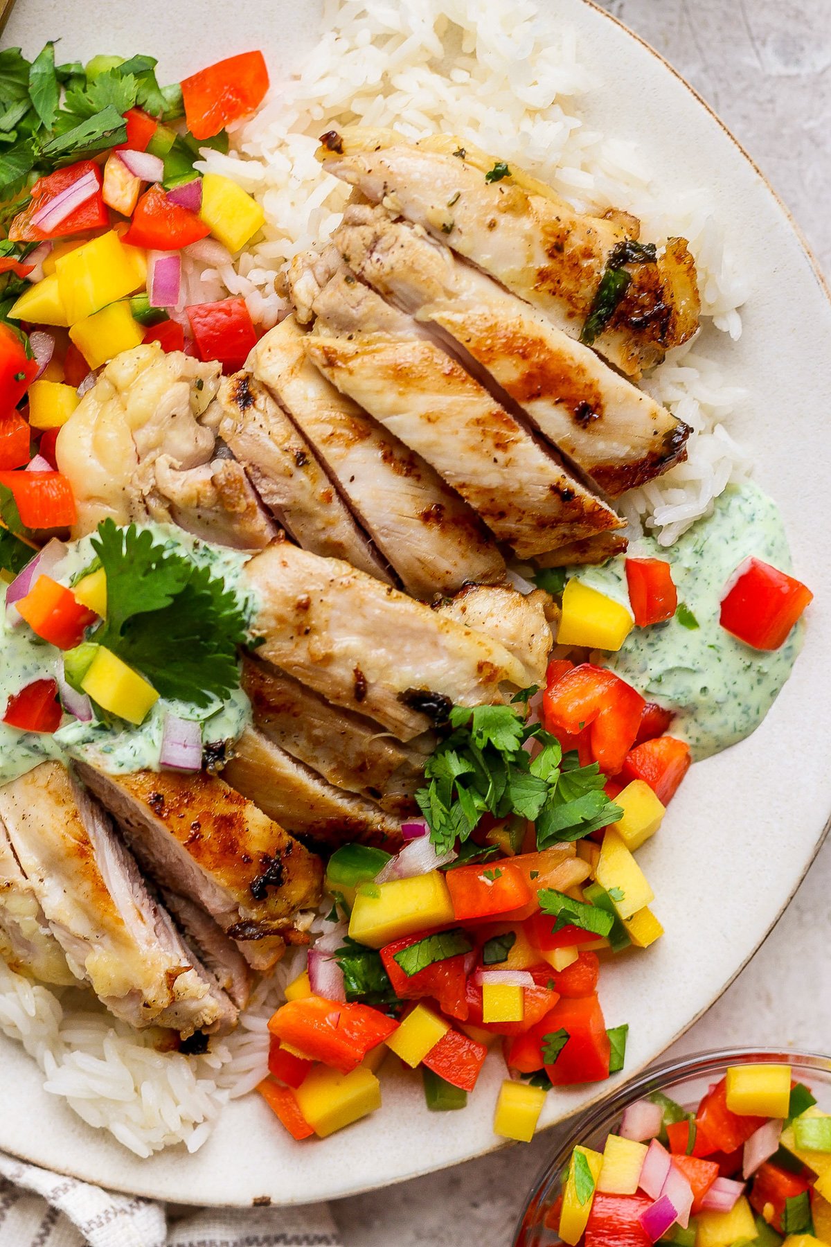 A chicken meal prep favorite: grilled chicken slices on white rice, topped with chopped mango salsa, fresh cilantro, and a dollop of creamy herb sauce.