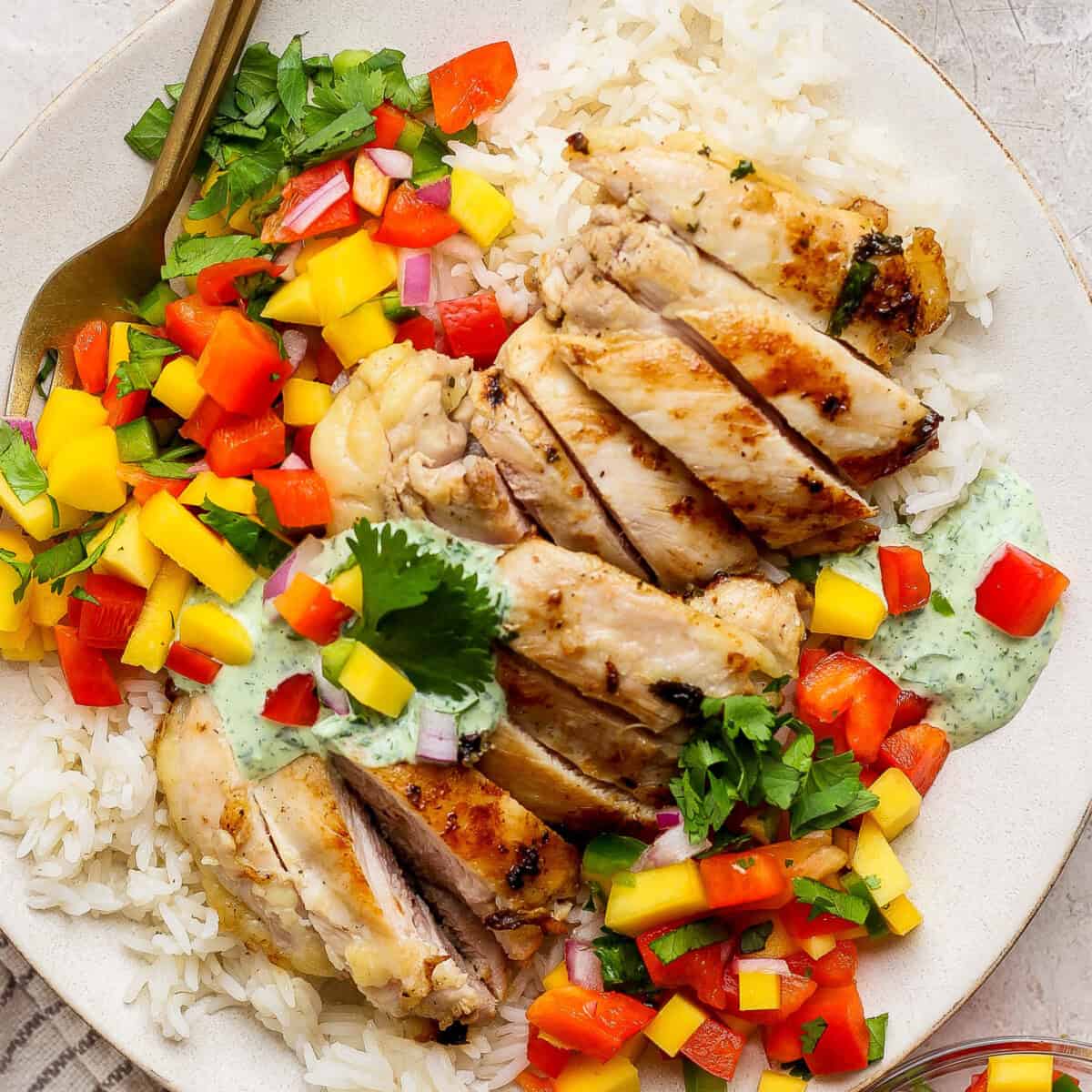A chicken meal prep plate featuring grilled chicken slices on white rice, topped with diced mango, red bell pepper, cilantro, and a drizzle of green sauce, served with a fork on the side.