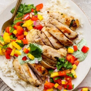 A chicken meal prep plate featuring grilled chicken slices on white rice, topped with diced mango, red bell pepper, cilantro, and a drizzle of green sauce, served with a fork on the side.