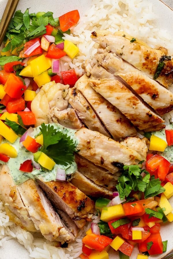 A chicken meal prep plate featuring grilled chicken slices on white rice, topped with diced mango, red bell pepper, cilantro, and a drizzle of green sauce, served with a fork on the side.