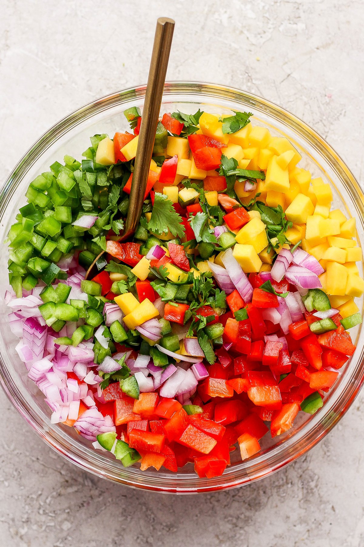A glass bowl filled with chopped red onion, red and green bell pepper, mango, jalapeño, and cilantro sits with a gold spoon on a light surface—perfect for adding freshness to your next chicken meal prep.