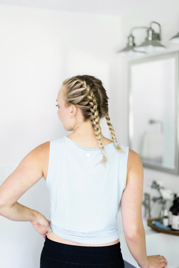 A woman demonstrating how to French braid her own hair in front of a mirror.