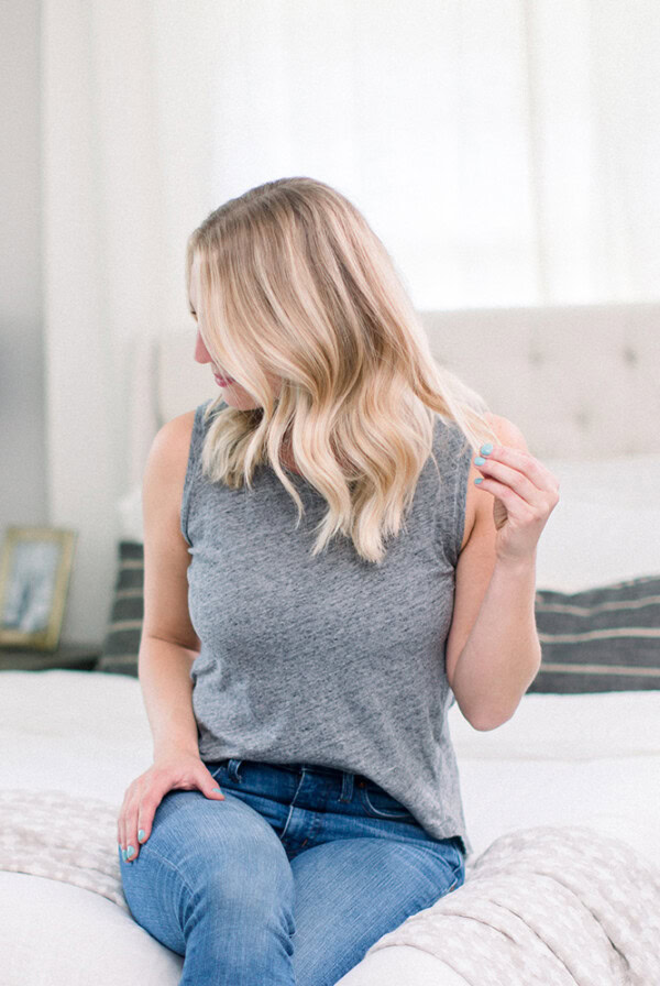 A blonde woman wearing jeans and a grey tank top demonstrates a loose wave hair tutorial while sitting on a bed.