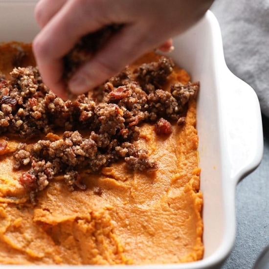 A person preparing a healthy sweet potato casserole by scooping mashed sweet potatoes into a dish.