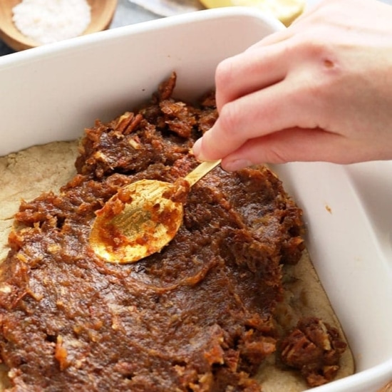 A person effortlessly prepares easy pecan pie bars by pouring a spoonful of pecans into a baking dish.