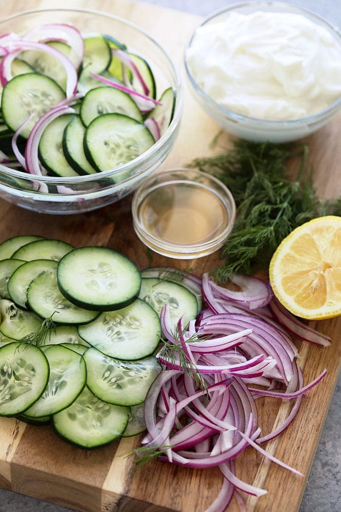 Cucumber, red onion, apple cider vinegar, Greek yogurt, fresh dill, and lemon on a cutting board. 