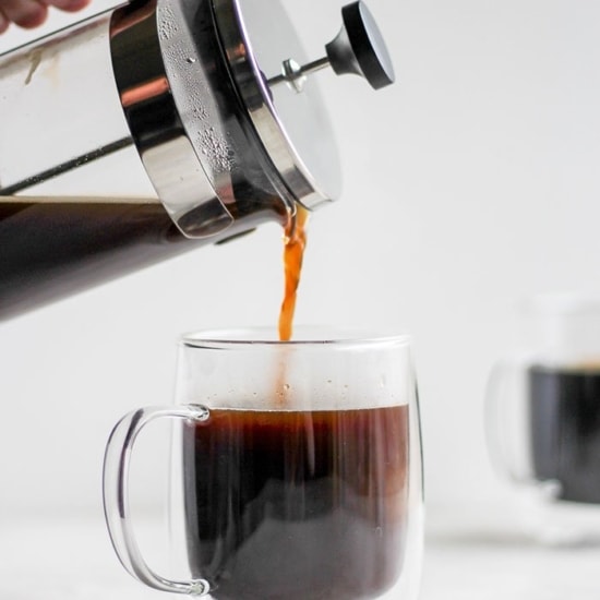 A person using a French press to pour coffee into a glass mug.