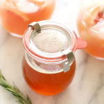 A glass jar with orange juice, honey simple syrup, and a rosemary sprig.