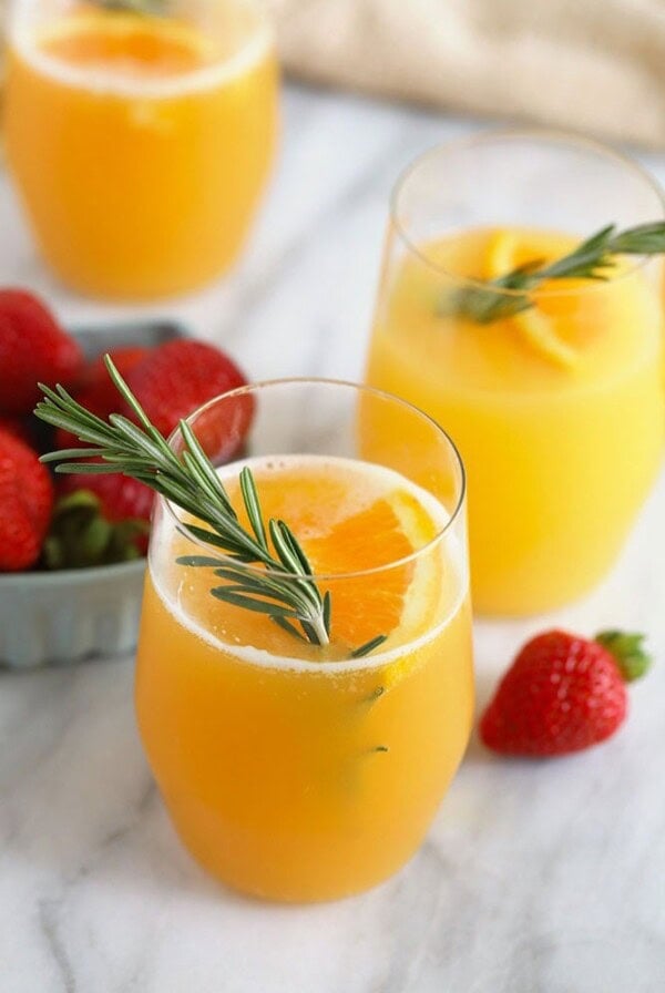 A refreshing twist on the classic beermosa, with two glasses of orange juice adorned with strawberries and sprigs of rosemary.