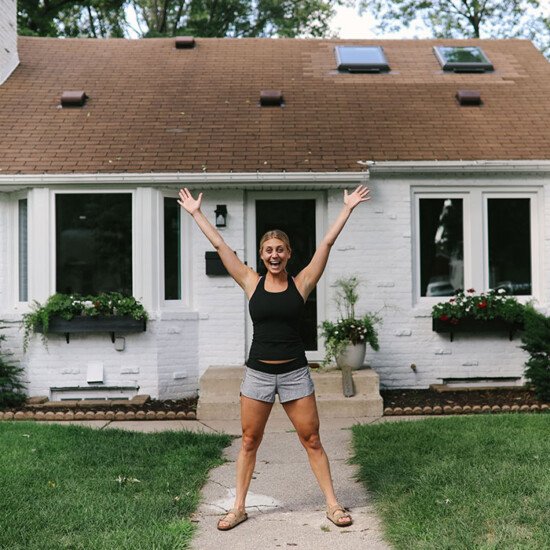 A woman standing in front of a white painted brick house with her arms outstretched.