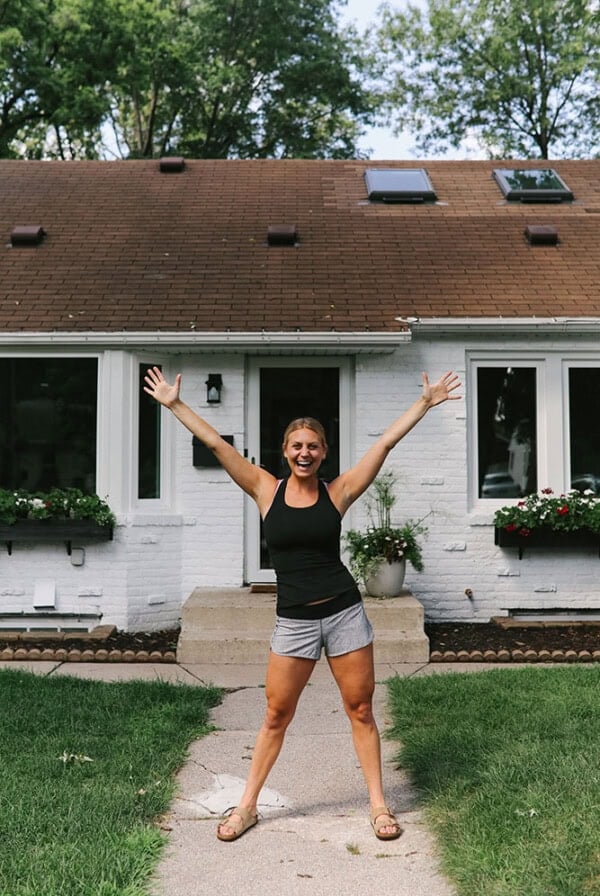 A woman posing with open arms in front of a white painted brick house.