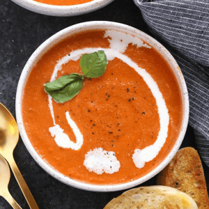 A bowl of creamy, Healthy Tomato Bisque Soup garnished with a dollop of cream and fresh basil, accompanied by slices of bread and golden spoons on the side.