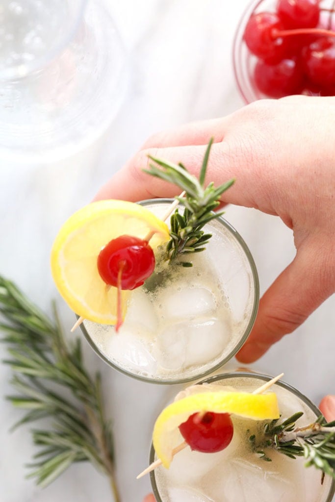 a person holding a glass of vodka collins with cherries and rosemary.