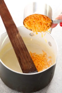 A person preparing cauliflower mac and cheese by pouring cheese into a pot with a wooden spoon.