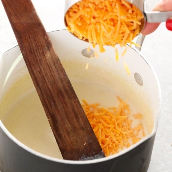 A person preparing cauliflower mac and cheese by pouring cheese into a pot with a wooden spoon.