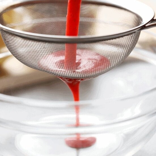 Blended strawberries being poured into a sieve over a glass bowl.