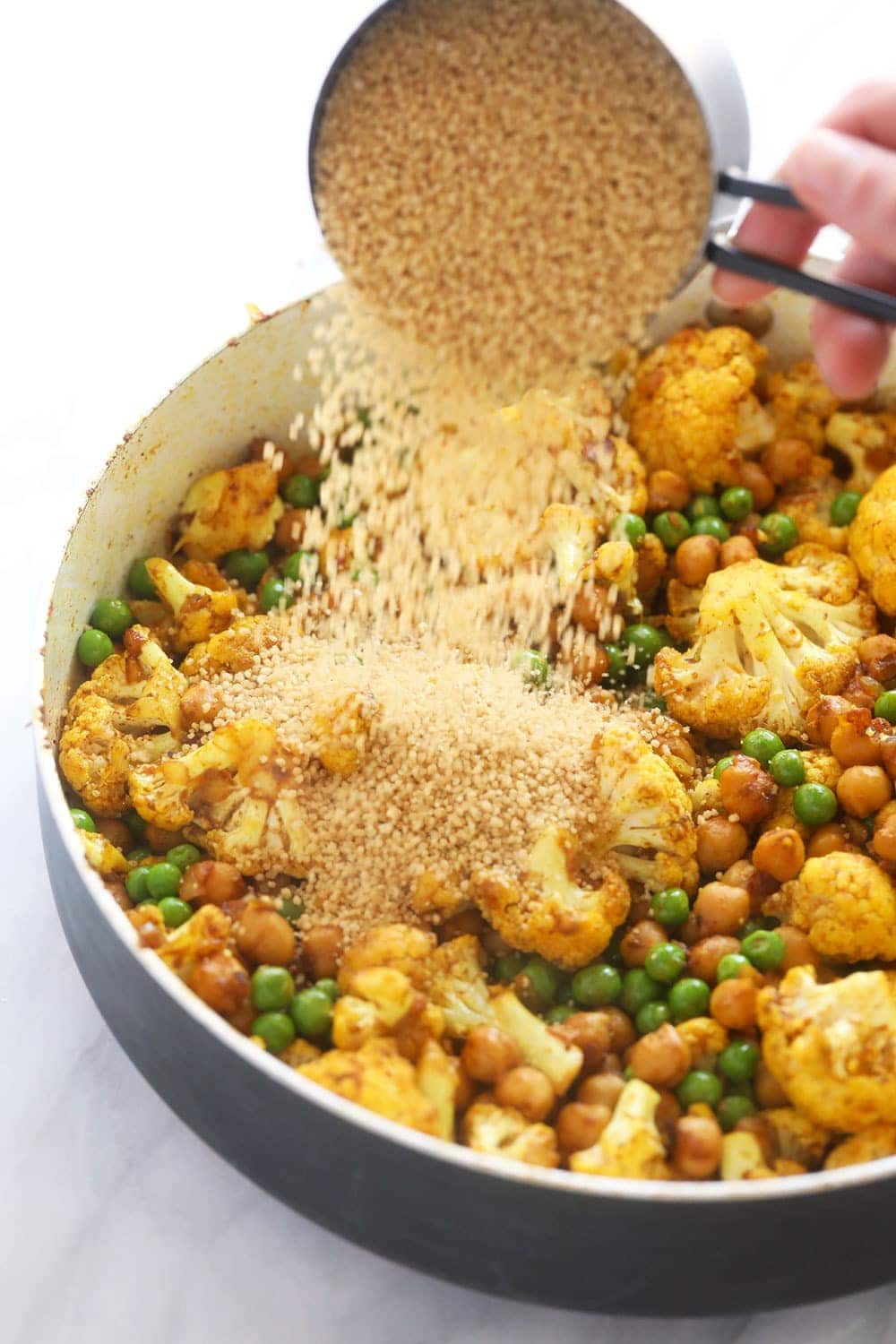 couscous being poured over the rest of the moroccan chickpea ingredients in a skillet