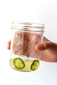 A hand holding a jar of tequila with jalapeño slices in it.