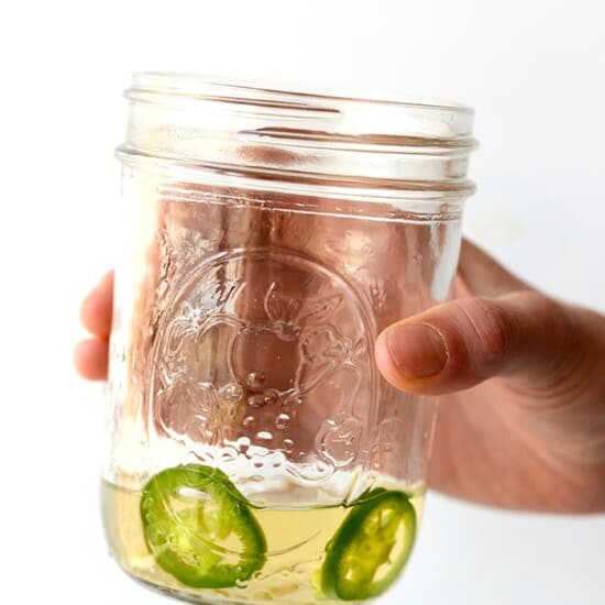 A hand holding a jar of tequila with jalapeño slices in it.