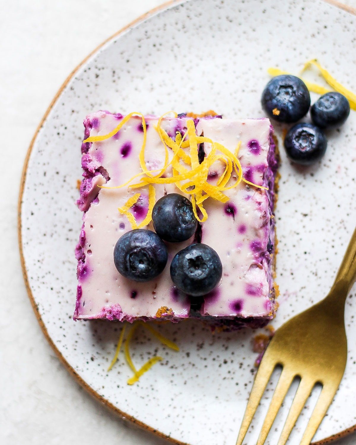 blueberry cheesecake bar on a plate topped with fresh blueberries and lemon zest