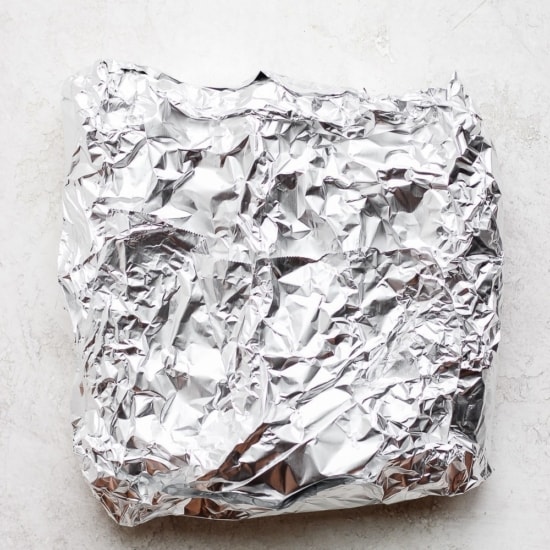 Grilled corn wrapped in aluminum foil on a white surface.