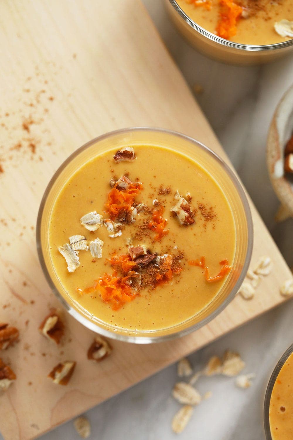 A glass with carrot cake smoothie topped with walnuts, oats, and shredded carrot. 