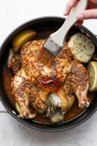 A person is roasting a whole chicken in a pan with lemons and rosemary.