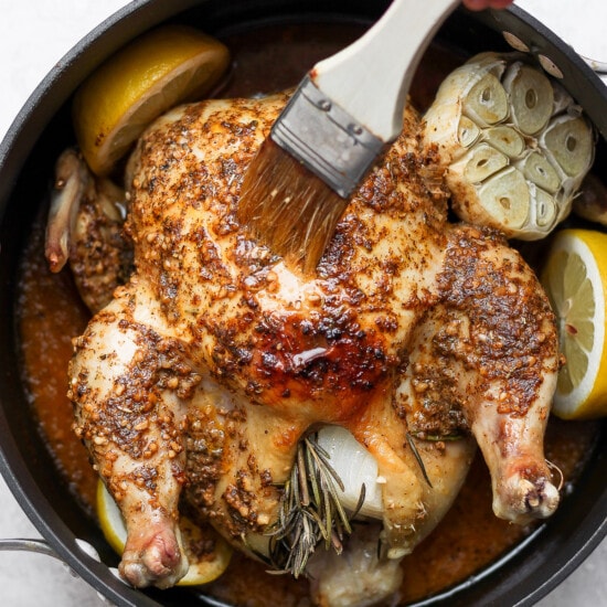 A person is roasting a whole chicken in a pan with lemons and rosemary.