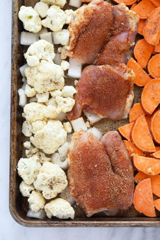 Chicken thighs, sweet potatoes, cauliflower, and onion on a baking sheet. 