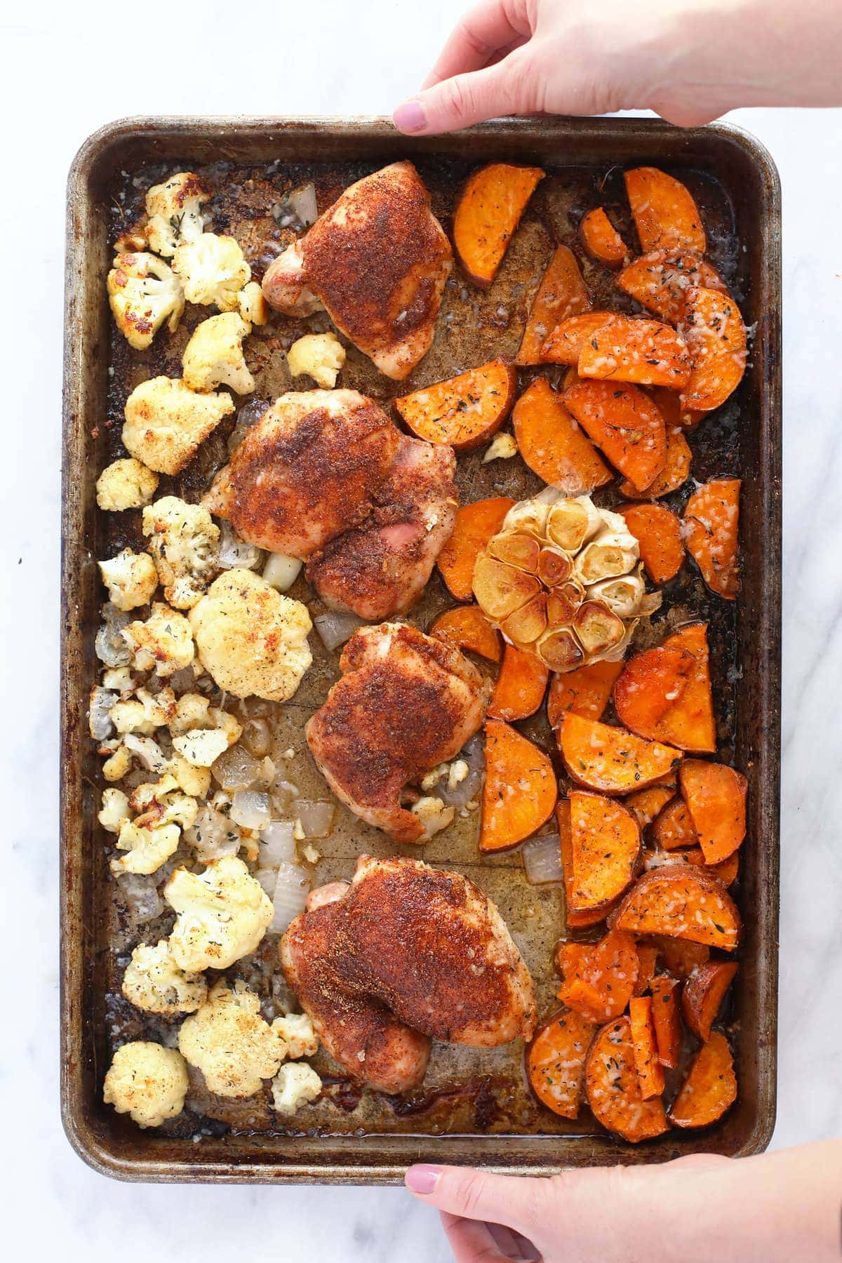 Chicken thighs, sweet potatoes, cauliflower, and garlic on a baking sheet. 