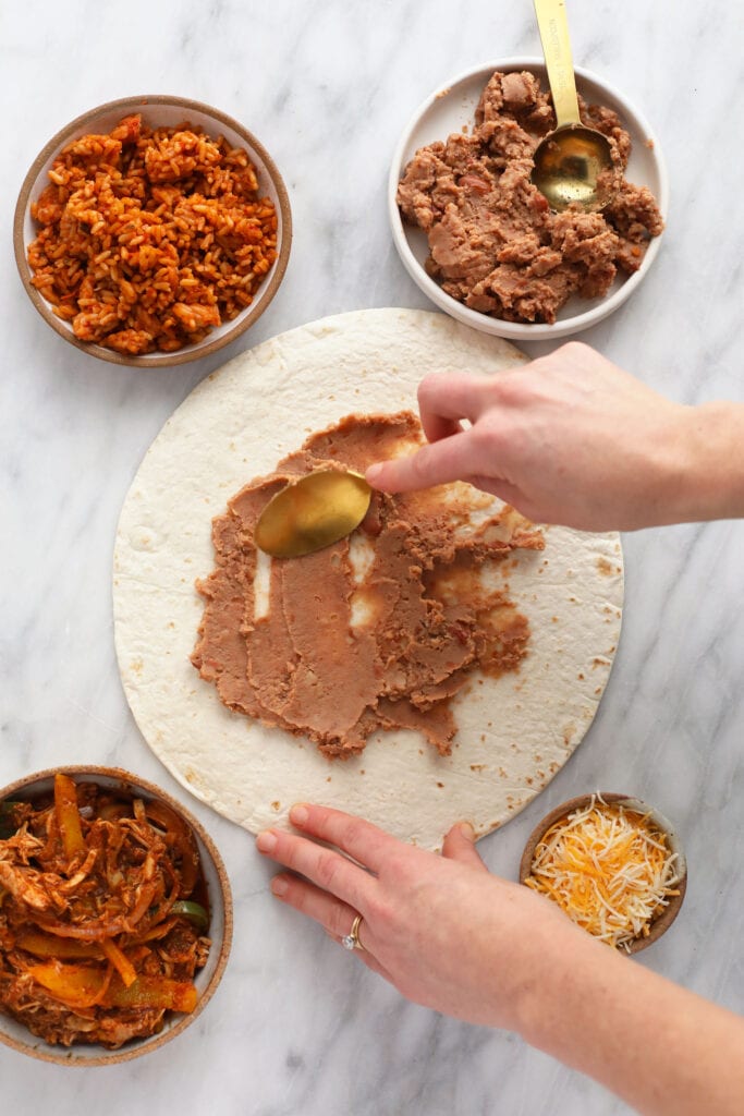 refried beans being spread on a tortilla for shredded chicken burritos.