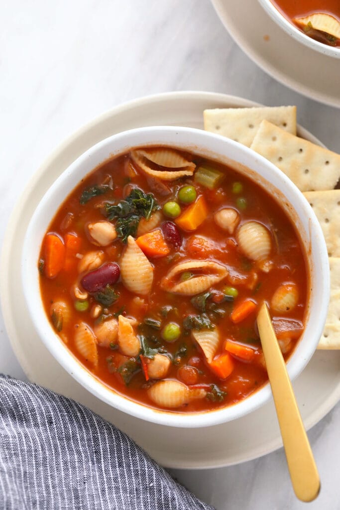 A bowl of minestrone soup with crackers on the plate beside it. 