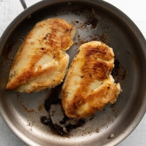 Two cooked chicken breasts in a stainless steel skillet. The chicken has a golden-brown sear and the pan shows some residual cooking marks.